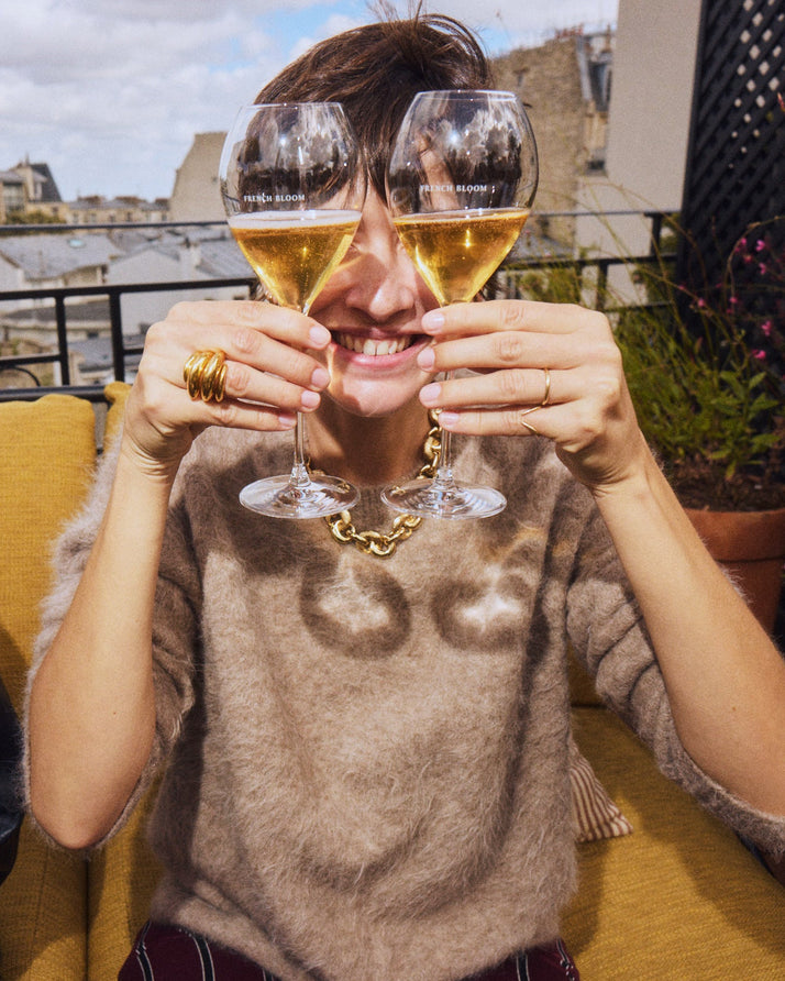 A woman on a rooftop terrace holds two wine glasses in front of her eyes, creating a playful mirror effect. She is wearing a fuzzy taupe-brown sweater with a dark brown graphic print, gold jewelry, and a gold chain necklace.
