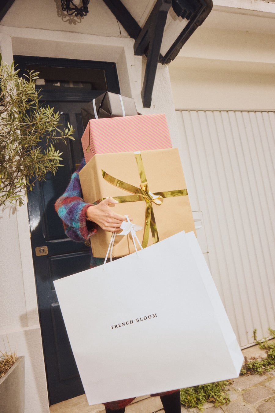 A woman carrying wrapped gifts and a French Bloom bag to a front door