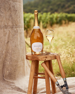 A bottle of white wine and a filled wine glass sit on a small wooden stool beside a large stone barrel, with a vineyard and hills blurred in the background. A pruning tool rests on the ground nearby.
