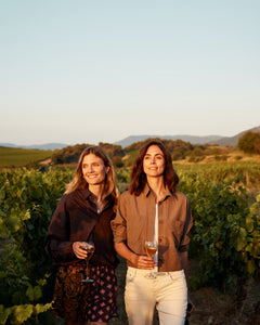 Two women standing in a vineyard at sunset, each holding a glass of white wine. The woman on the left wears a dark brown jacket and a dark patterned skirt, while the woman on the right wears a brown button-up shirt and light beige pants.