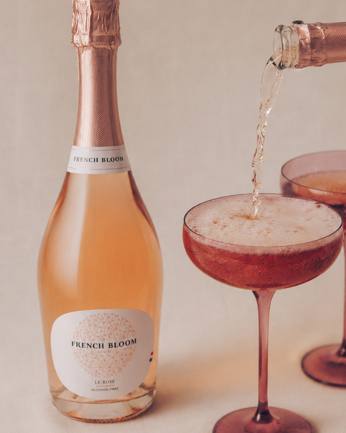 A bottle of French Bloom Rosé  champagne stands next to a bottle being poured into two coupe glasses on a beige fabric background.