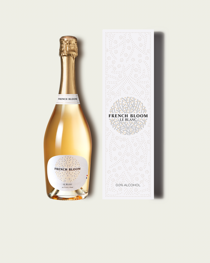 A bottle of French Bloom Le Blanc sparkling wine with pale golden liquid, shown next to its tall white patterned box on a light beige background.