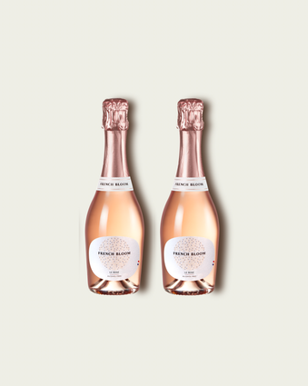 Two half bottles of French Bloom Le Rosé sparkling wine with pale pink liquid and metallic rose-gold foil tops, set against a light warm-gray background.