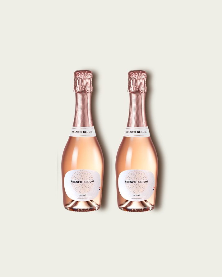 Two half bottles of French Bloom Le Rosé sparkling wine with pale pink liquid and metallic rose-gold foil tops, set against a light warm-gray background.
