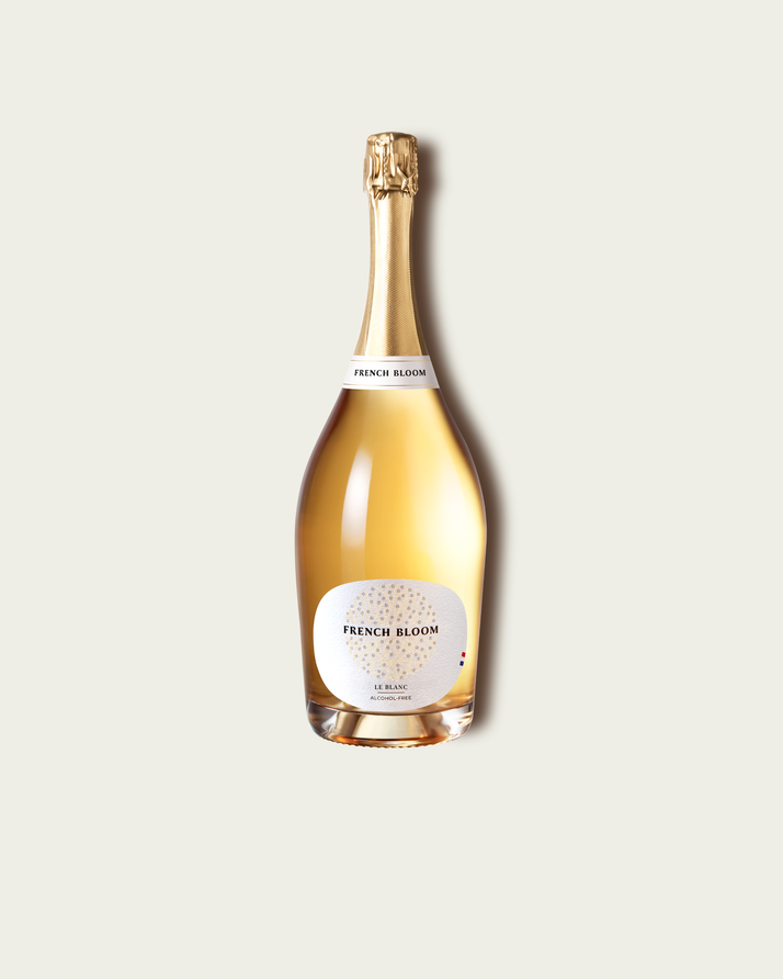 A magnum bottle of French Bloom Le Blanc sparkling wine with pale golden liquid and a metallic gold foil top, set against a light warm-gray background.