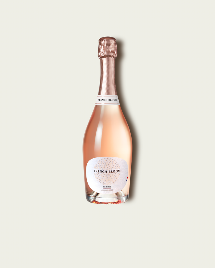 A bottle of French Bloom Le Rosé with pale pink liquid, a white label, and a metallic rose gold foil top, centered on a light beige background.