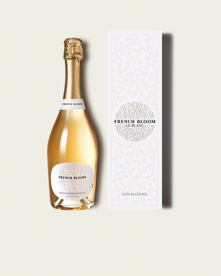 A bottle of French Bloom Blanc non-alcoholic sparkling wine with gold foil and a white label, shown next to its tall white patterned box on a light beige background.