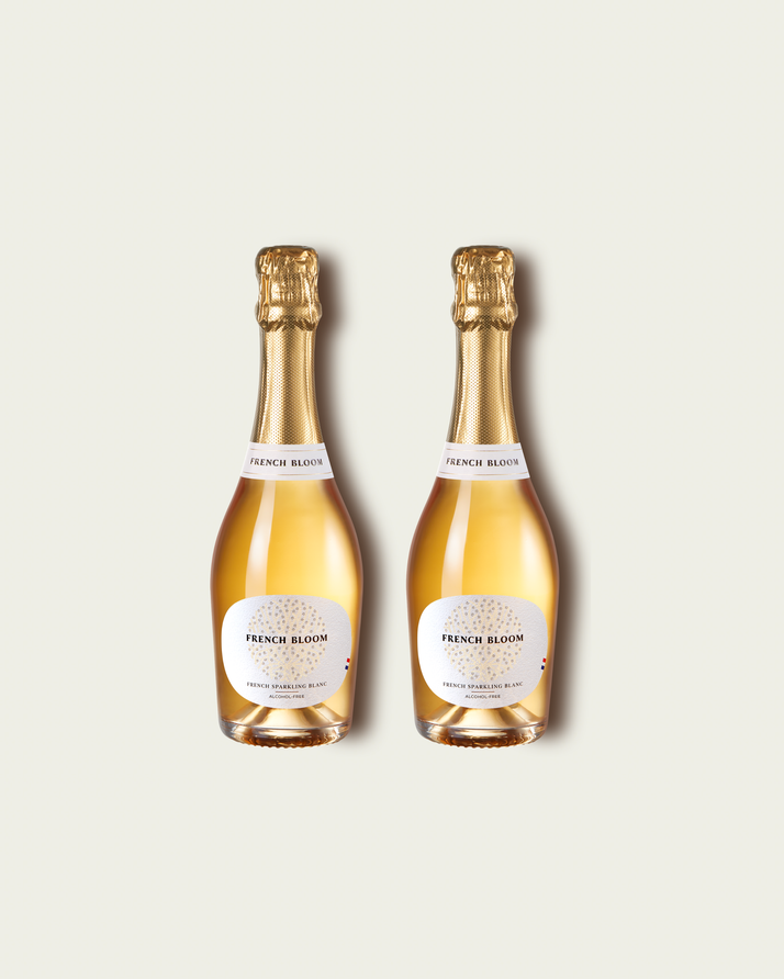 Two half bottles of French Bloom Le Blanc sparkling wine with pale golden liquid and metallic gold foil tops, set against a light warm-gray background.