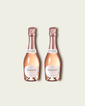 Two half bottles of French Bloom Le Rosé sparkling wine with pale pink liquid and metallic rose-gold foil tops, set against a light warm-gray background.