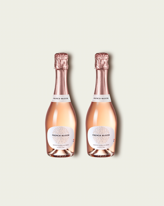 Two half bottles of French Bloom Le Rosé sparkling wine with pale pink liquid and metallic rose-gold foil tops, set against a light warm-gray background.