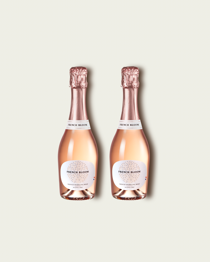 Two half bottles of French Bloom Le Rosé sparkling wine with pale pink liquid and metallic rose-gold foil tops, set against a light warm-gray background.
