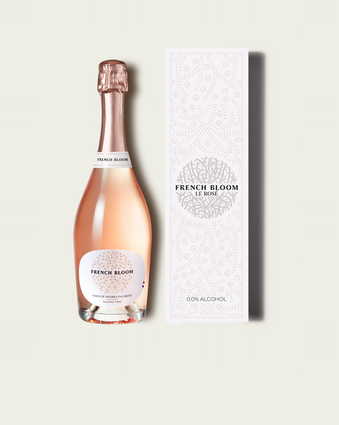 A bottle of French Bloom sparkling wine with pale pink liquid and a metallic rose-gold foil top, shown next to its tall white patterned box on a light warm-gray background.