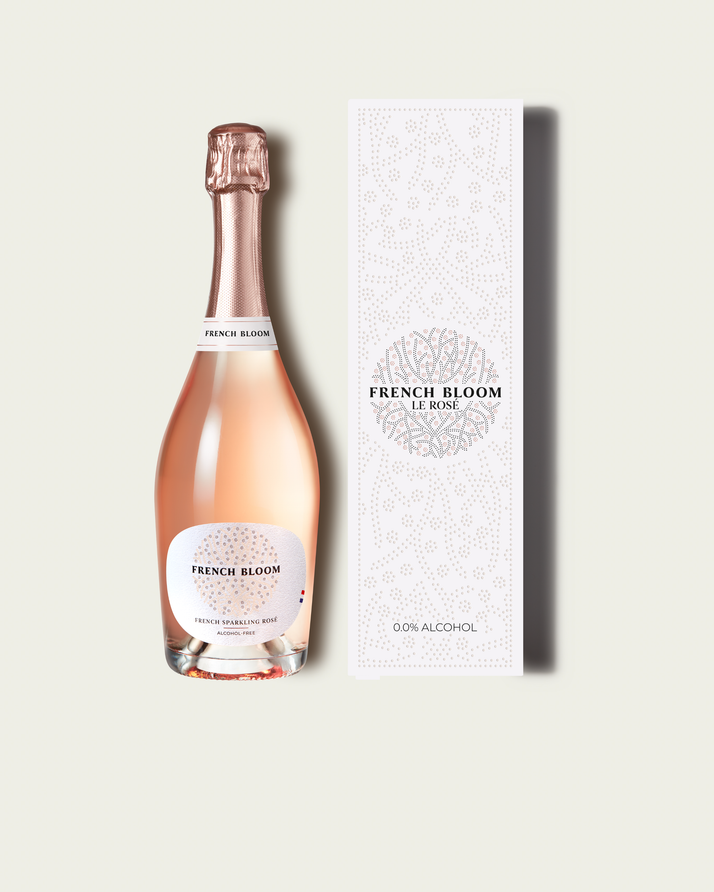 A bottle of French Bloom sparkling wine with pale pink liquid and a metallic rose-gold foil top, shown next to its tall white patterned box on a light warm-gray background.