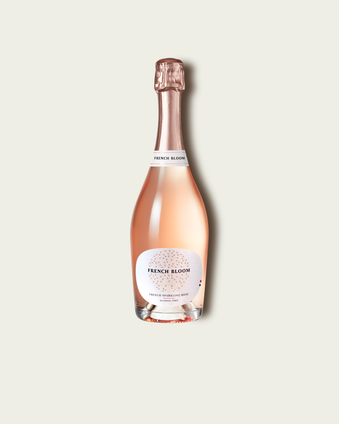 A bottle of French Bloom Le Rosé with pale pink liquid, a white label, and a metallic rose gold foil top, centered on a light beige background.