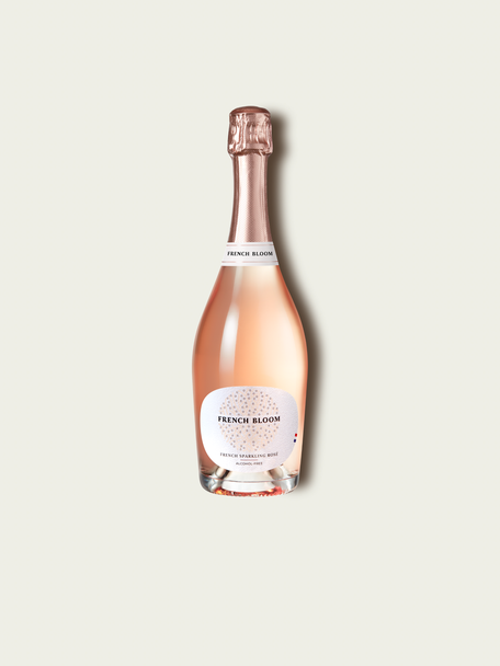 A bottle of French Bloom Le Rosé with pale pink liquid, a white label, and a metallic rose gold foil top, centered on a light beige background.
