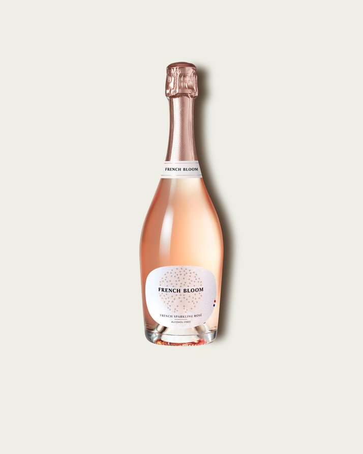 A bottle of French Bloom Le Rosé with pale pink liquid, a white label, and a metallic rose gold foil top, centered on a light beige background.