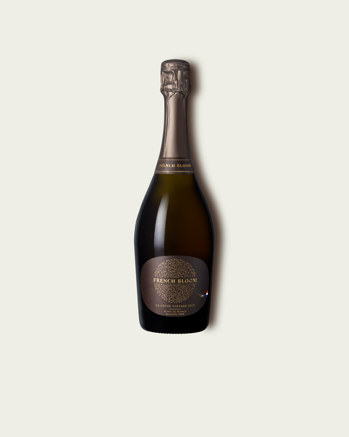A bottle of French Blanquette de Champagne with a dark brown glass body, gold foil, and a brown label with gold lettering, centered on a light beige background.