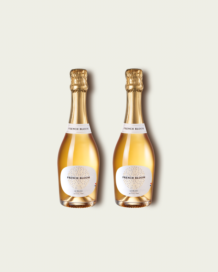 Two half bottles of French Bloom Le Blanc sparkling wine with pale golden liquid and metallic gold foil tops, set against a light warm-gray background.