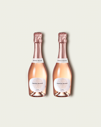 Two half bottles of French Bloom Le Rosé sparkling wine with pale pink liquid and metallic rose-gold foil tops, set against a light warm-gray background.