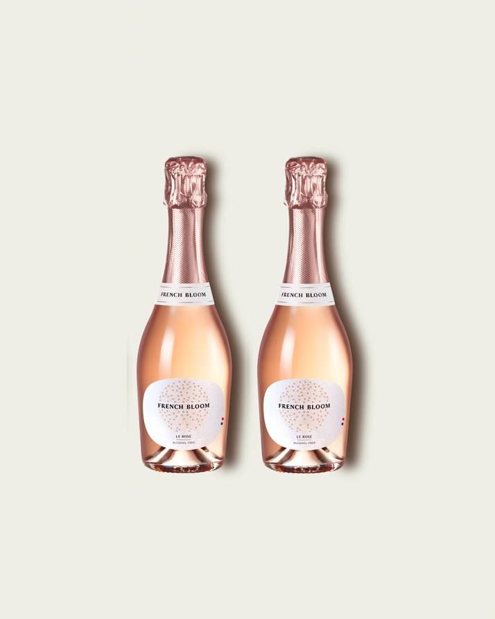 Two half bottles of French Bloom Le Rosé sparkling wine with pale pink liquid and metallic rose-gold foil tops, set against a light warm-gray background.