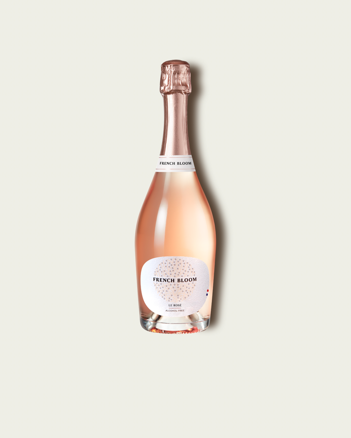 A bottle of French Bloom Le Rosé with pale pink liquid, a white label, and a metallic rose gold foil top, centered on a light beige background.