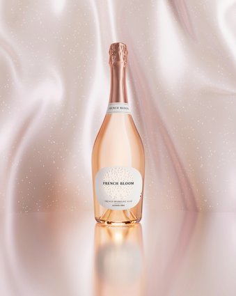 A bottle of French Bloom Le Rosé non alcoholic sparkling wine with pale pink liquid and a metallic rose-gold foil top, set against a softly draped blush satin background with subtle sparkling details and a reflective surface.
