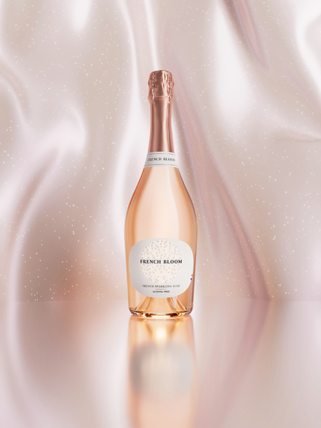 A bottle of French Bloom Le Rosé non alcoholic sparkling wine with pale pink liquid and a metallic rose-gold foil top, set against a softly draped blush satin background with subtle sparkling details and a reflective surface.