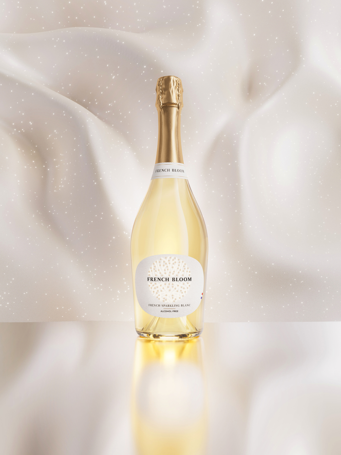 A bottle of French Bloom Non Alcoholic  with a gold foil top and a white label, set against a soft, draped white fabric background with subtle sparkles.